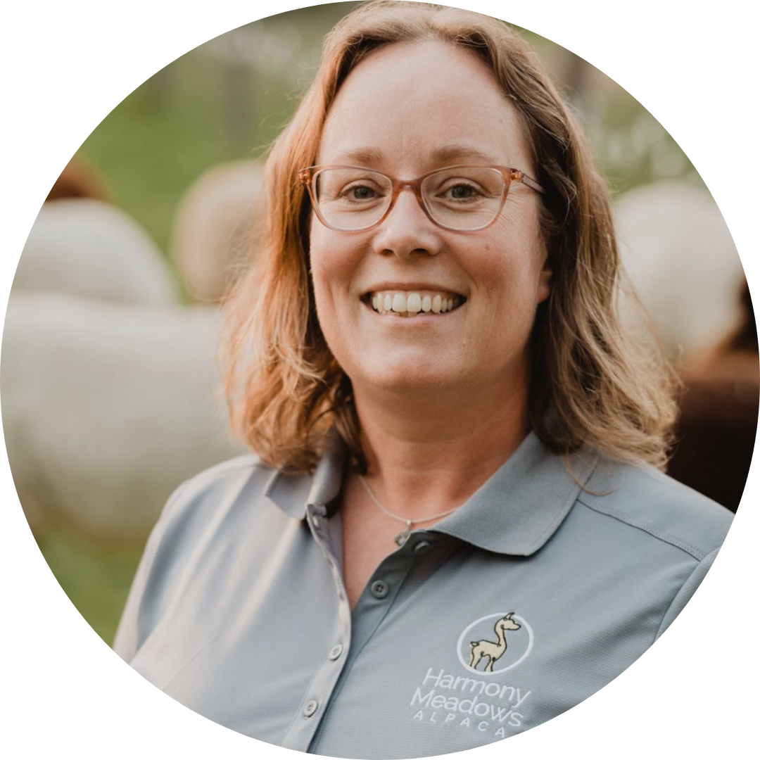 Pictured is Kristi Mercier, owner of Harmony Meadows Alpaca Farm. She's wearing round glasses and smiling at the camera in her grey Harmony Meadows shirt, which has the business logo on it.